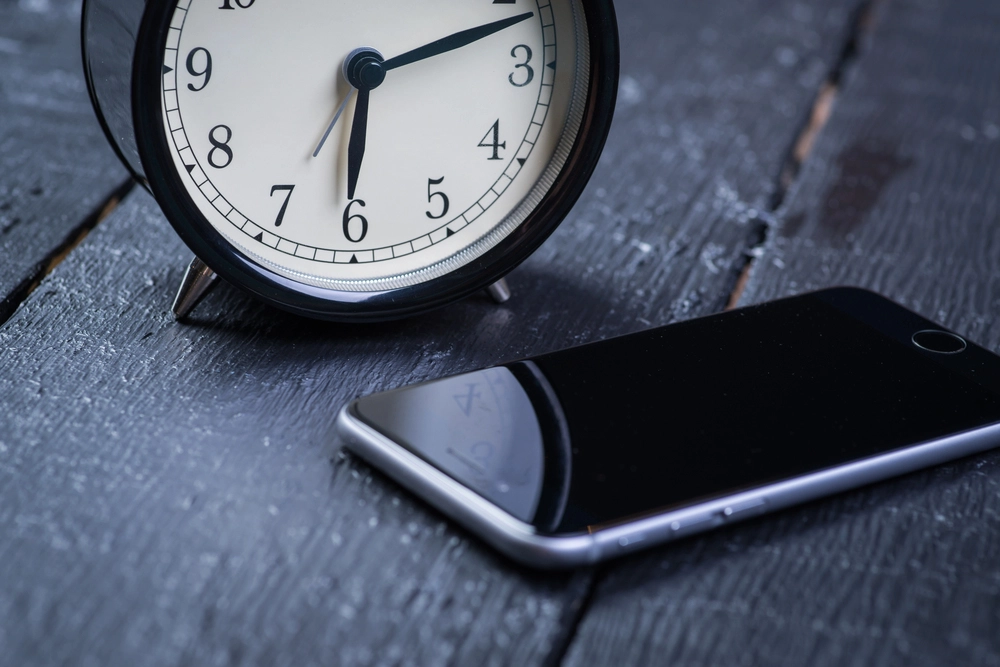 image of a phone with a black screen next to a clock on a dark wood tabletop